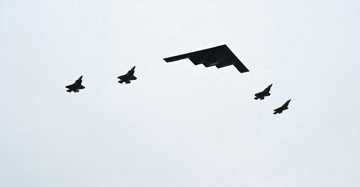 A B-2 bomber flanked by four F-25 fighters fly overhead against an overcast sky.