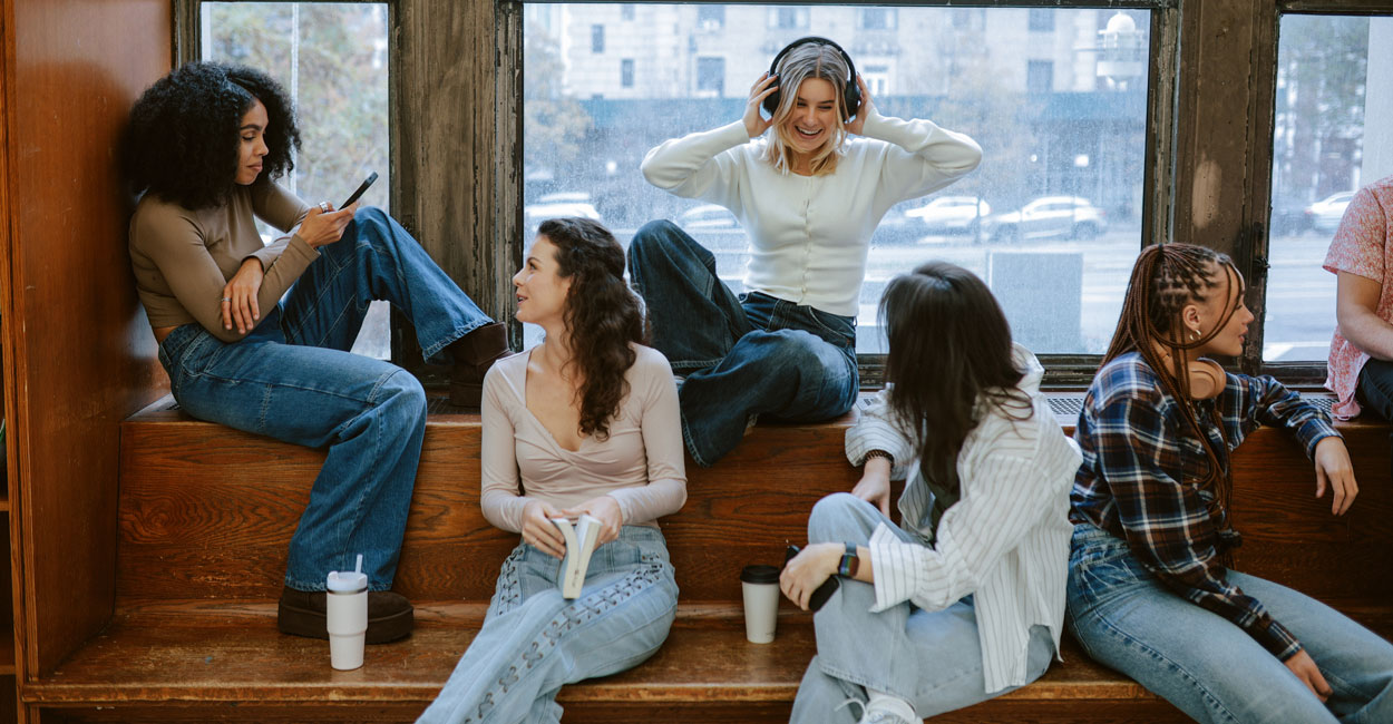 A mixed-raced group of young women sit on a bench and ledge interacting.
