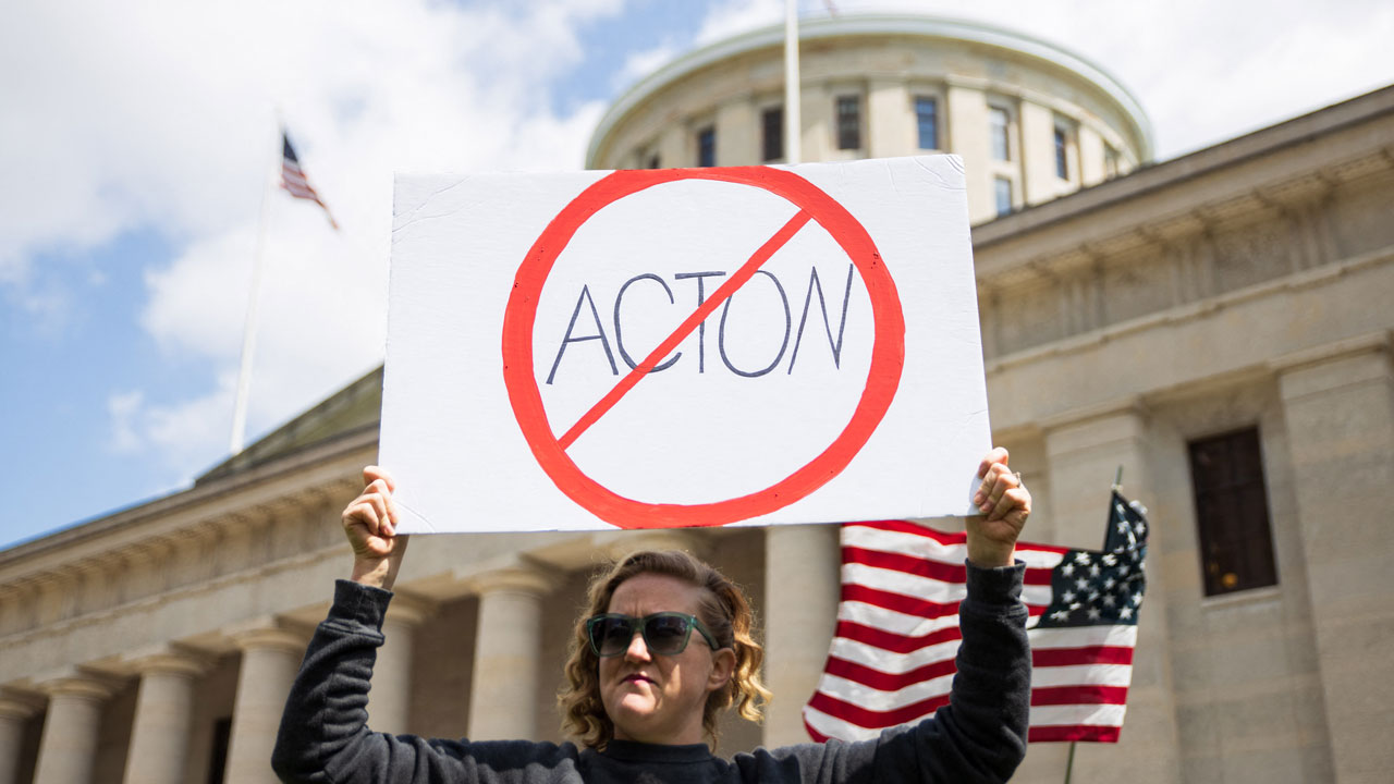 A woman stands outside of the Ohio capitol with a prohibition slash through symbol sign of "Acton."